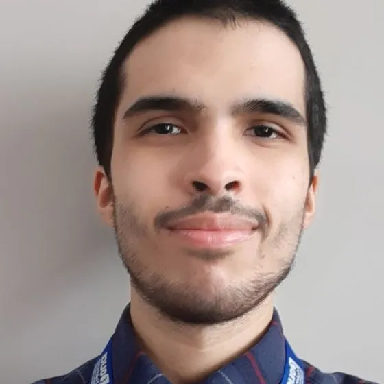 A young man with short black hair and light facial hair, wearing a blue plaid shirt with red stripes. He is facing forwards with a slight smile.